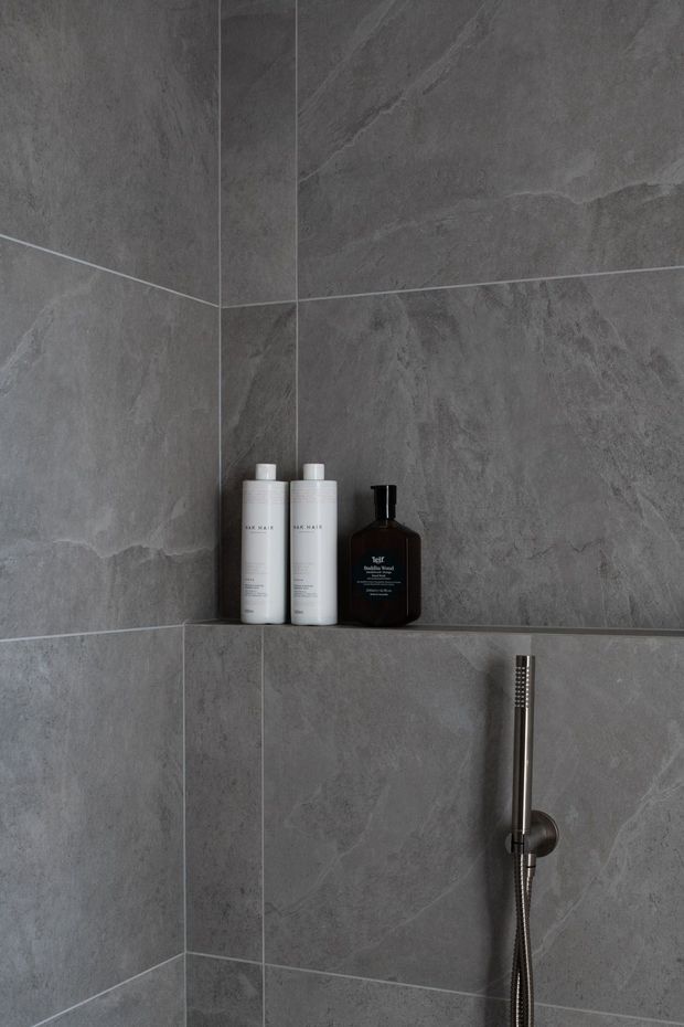 Close-up of a minimalist shower enclosure corner clad in large-format dark grey stone-look tiles. A recessed wall niche holds shampoo and soap bottles, while a slim brushed metal handheld shower fixture is mounted below. The image has a restrained, monochromatic and contemporary appearance.