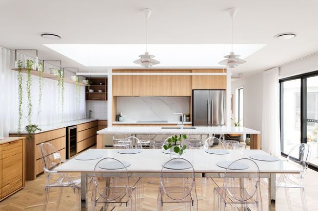 Bright open-plan kitchen and dining area with extensive light timber cabinetry, a large white island and dining table, transparent acrylic dining chairs, stainless-steel appliances, hanging plants, and large glazed doors and windows. A broad skylight and white finishes create an airy contemporary interior.