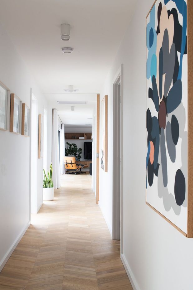 A bright, narrow residential hallway with white walls and ceiling, pale timber herringbone flooring, multiple white doorways, and framed abstract artwork along the walls. The corridor leads toward an open living area with a timber-toned built-in media unit, indoor plant, and mid-century-style lounge chair visible at the far end. Recessed and surface-mounted ceiling lights provide even illumination.