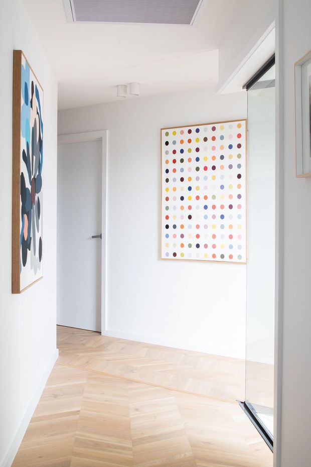 Bright, minimalist hallway or landing with white walls, pale timber herringbone parquet flooring, frameless or slim-framed internal glazing, and colorful framed abstract artwork. A white flush door and recessed ceiling air-conditioning grille are visible, with strong natural light entering from the glazed opening on the right.