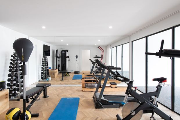Bright, modern home gym with extensive cardio and strength-training equipment, dumbbell racks, exercise mats, mirrors, and full-height glazing overlooking water. The room has a white ceiling and walls, pale herringbone timber flooring, and a clean open layout.