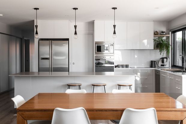 Bright contemporary open-plan kitchen and dining area with extensive glossy white cabinetry, integrated stainless-steel appliances, a large central island with seating, and a substantial warm timber dining table. Minimal pendant lights, dark flooring, and large glazed doors/windows create a clean, refined interior.