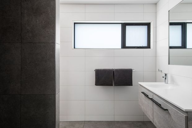 A contemporary bathroom with large white wall tiles, a dark tiled shower or feature wall at left, a floating light-grey vanity with a white benchtop, chrome tapware, wall-mounted towel rail, black-framed frosted windows, and a large mirror. The room has a clean, restrained palette of white, grey, and black.