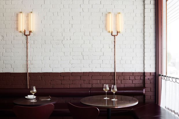 A stylish restaurant or bar seating area with built-in dark burgundy banquette seating, round marble-topped tables, and curved burgundy chairs. The two-tone exposed brick wall has a white upper section and deep red lower section, complemented by slim brass-and-glass wall sconces. A large window with sheer curtains provides natural light. The space has a refined, moody, Art Deco-inspired atmosphere.