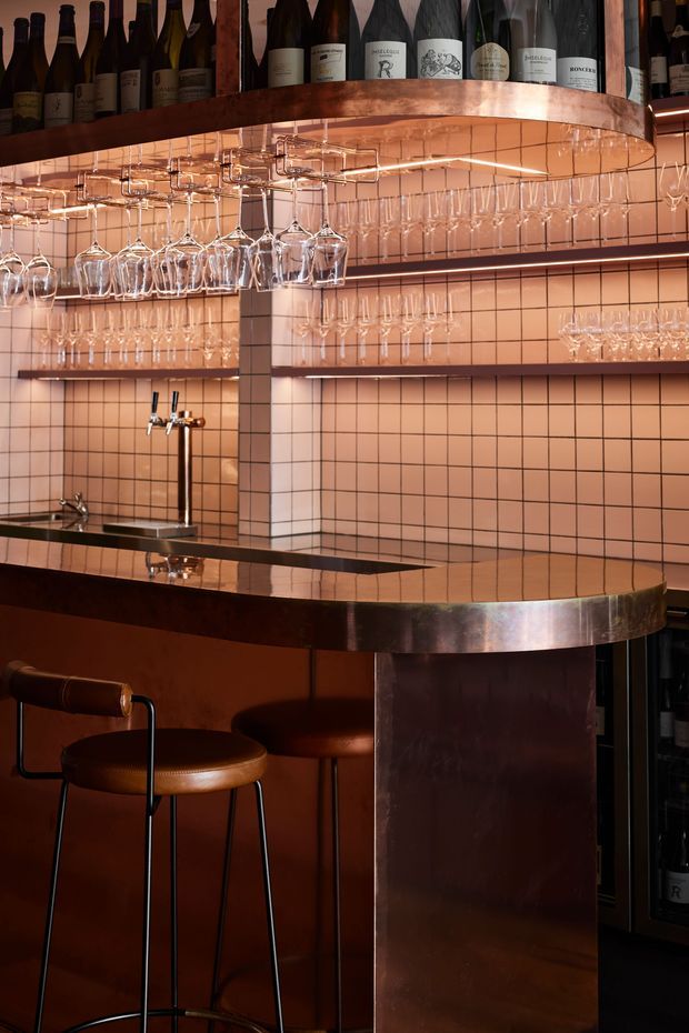 A warmly lit, compact bar interior with a polished dark counter, copper-toned metal edging and cabinetry, peach-pink square tiled walls, extensive suspended wine and cocktail glass storage, and wine bottles displayed on an overhead shelf. Brown leather bar stools and beer taps reinforce the intimate hospitality setting. The composition has a refined, moody, richly coloured contemporary feel.