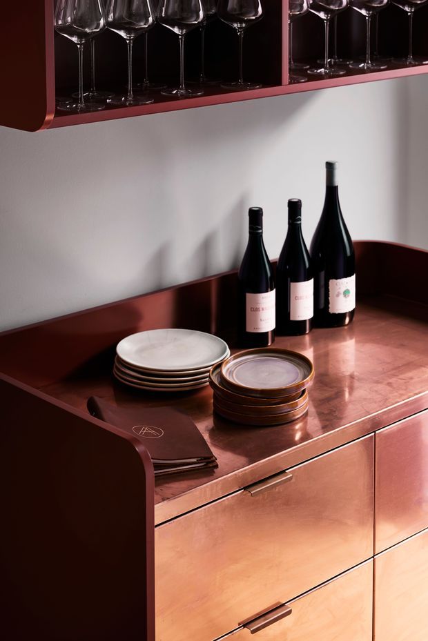A close-up of a refined wine and serving station with dark copper-toned cabinetry and countertop, curved side panels, open glassware shelving, wine bottles, stacked ceramic plates, and leather-bound menus. The composition has warm dramatic lighting and a polished, luxurious hospitality-inspired feel.