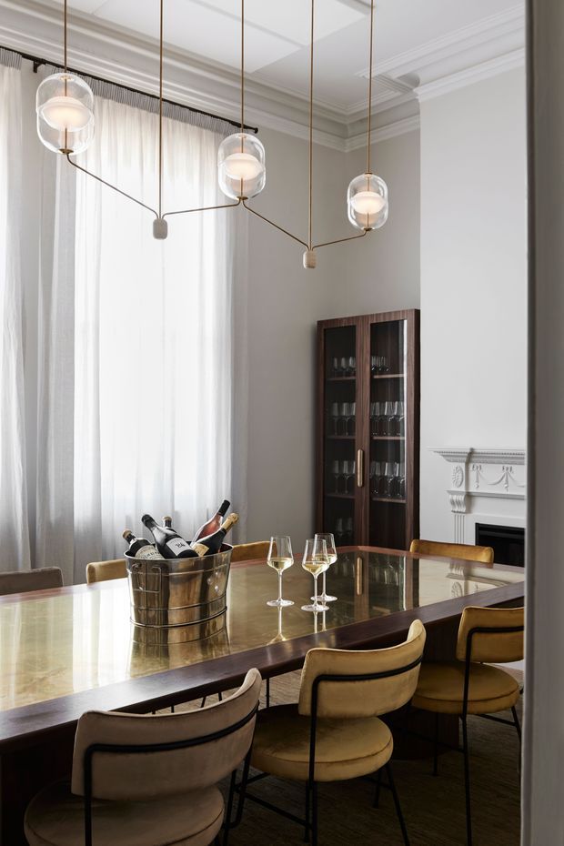 Elegant formal dining room with a long reflective dining table surrounded by upholstered dining chairs. The room features tall sheer curtains, ornate ceiling cornices and wall mouldings, a glass-fronted dark timber drinks cabinet, a decorative fireplace surround, and sculptural suspended pendant lights with brass-toned frames. The palette is predominantly warm neutral, white, taupe, dark timber, and metallic brass.