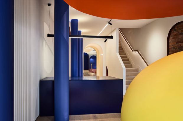 A bold, sculptural interior entry or circulation space featuring deep blue cylindrical columns and a dark blue built-in counter or partition. Curved orange and yellow ceiling or sculptural elements frame the view, while a staircase rises at the rear beside a warmly lit arched opening and exposed brick feature wall. The space has strong geometric forms, dramatic colour blocking, integrated linear lighting, and a playful contemporary character.