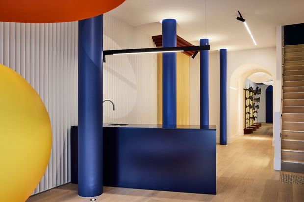 A bold, colourful contemporary interior featuring a dark blue reception or kitchen-like counter, multiple cylindrical blue columns, bright yellow and orange curved ceiling or wall elements, white fluted wall panelling, and light timber flooring. An arched hallway leads deeper into the space, with a staircase visible on the right and built-in shelving or display joinery beyond.