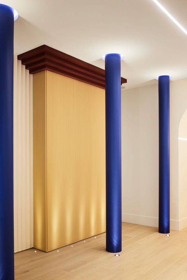 Interior architectural view featuring a minimalist gallery-like space with a warm pale timber floor, cream walls and ceiling, and several vivid cobalt-blue cylindrical columns. A tall ribbed golden-yellow feature wall is framed by layered dark reddish-brown horizontal timber beams, while slim linear ceiling lighting and small recessed floor lights add emphasis. The composition is bold, geometric, and highly curated.