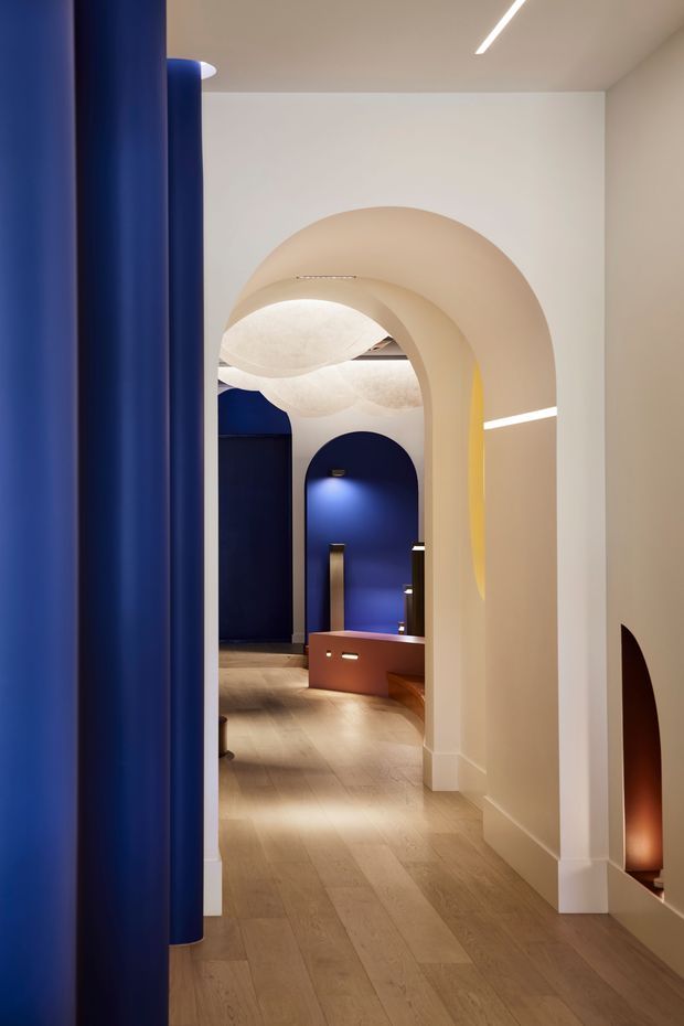 A refined interior hallway or entry passage with a series of rounded archways and sculptural curved ceiling forms. The space combines warm pale timber flooring and off-white plaster walls with deep navy-blue curved wall panels and concealed linear lighting. A low terracotta-toned built-in bench or joinery element is visible in the background, while a narrow arched wall niche adds a warm illuminated accent. The design has a highly contemporary, sculptural character with strong geometric colour blocking.