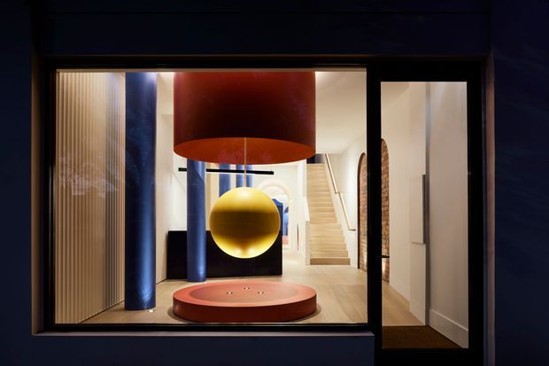 A dramatic contemporary interior viewed through a large dark-framed glazed opening. The entry or living space features a sculptural red circular platform, a suspended yellow spherical light beneath a large red cylindrical pendant, blue cylindrical columns, vertical timber screening, pale timber flooring, and a staircase leading upward in the background. The composition is bold, theatrical, and highly geometric.
