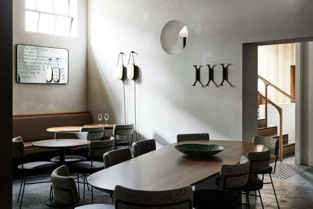 A refined contemporary dining and lounge area with a large oval dining table, upholstered dining chairs, a secondary round table, and a built-in banquette. Soft daylight enters through high windows, highlighting pale plaster walls, dark timber detailing, and sculptural wall-mounted lighting. A doorway opens toward a stairwell with timber handrails and vertical timber screening.