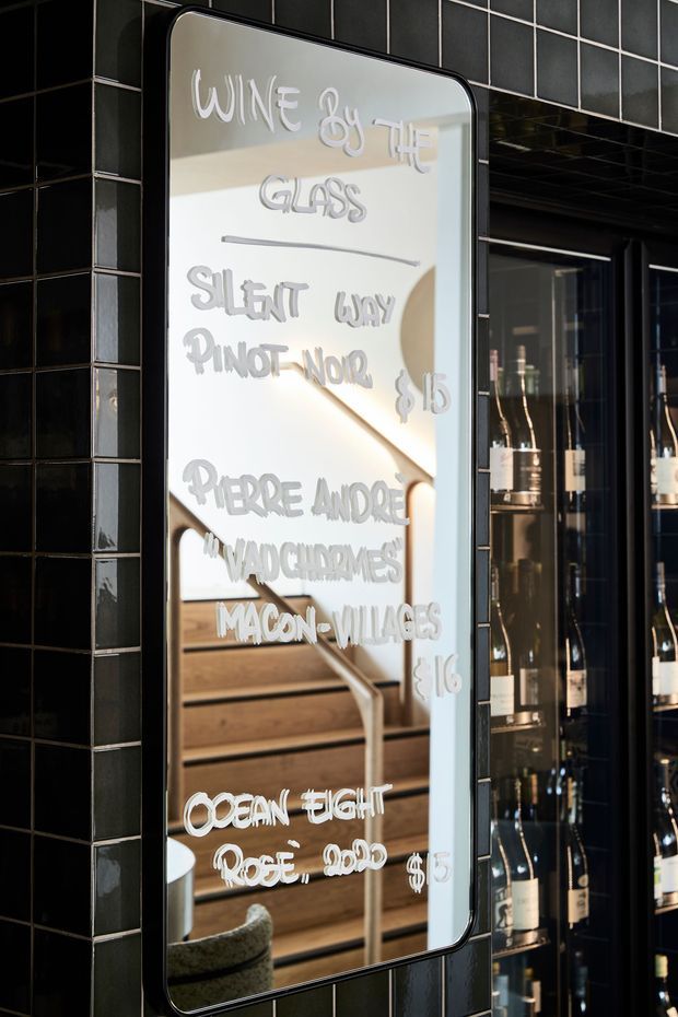 Close-up of a tall framed menu mirror mounted against a glossy black tiled wall. Handwritten white lettering lists wines and prices, while the reflection shows timber stair treads, a pale handrail, and part of the venue interior. Glass-front wine shelving with bottles is visible to the right, creating a refined hospitality setting with strong black, white, and warm timber tones.