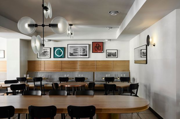 Contemporary restaurant or café dining interior with rows of small dark-framed tables and black chairs, a large rounded communal timber table in the foreground, and a long built-in banquette along the rear wall. The space features warm timber cabinetry and seating, a grey stone or concrete-look backsplash, white walls and ceiling, framed graphic artwork, and distinctive globe and disc wall lighting. The low ceiling and compact furniture arrangement create a refined, inviting hospitality setting.
