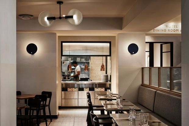 Interior view of a refined hotel dining room or restaurant, with neatly arranged dining tables and upholstered banquette seating in the foreground. A compact open kitchen with stainless-steel equipment and tiled surfaces is visible at the rear, separated by a black-framed glazed opening. Warm beige walls, dark timber or metal details, globe pendant lighting, and circular wall sconces create a polished contemporary hospitality setting. Signage indicates the hotel dining room.