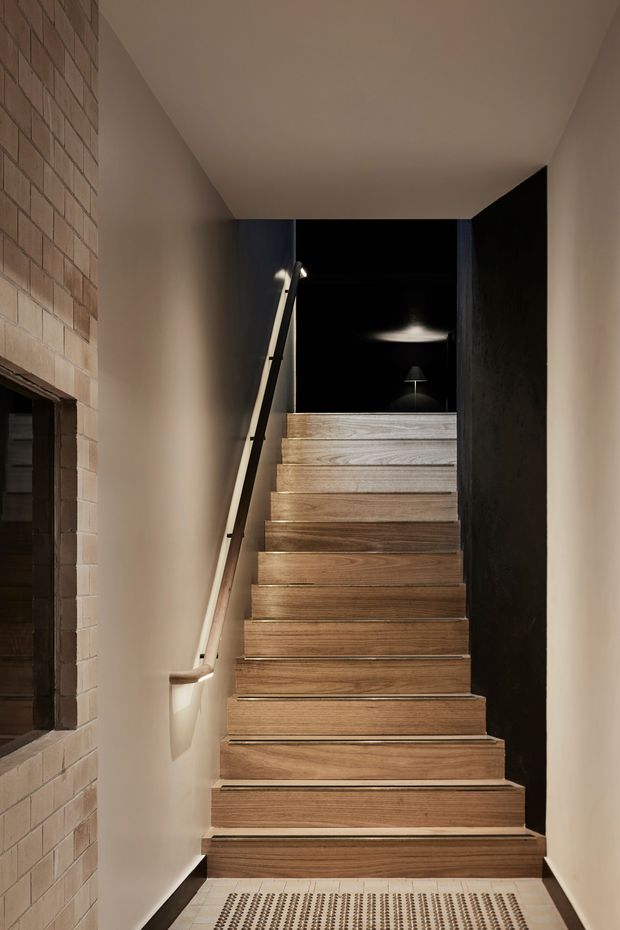 A narrow interior stairway leading upward, with warm timber-clad treads and risers, a simple pale handrail, smooth light-painted walls, and a dark feature wall at the upper landing. The left wall includes exposed light brickwork and a recessed opening. The stairwell is dramatically lit with a warm, refined contemporary feel.