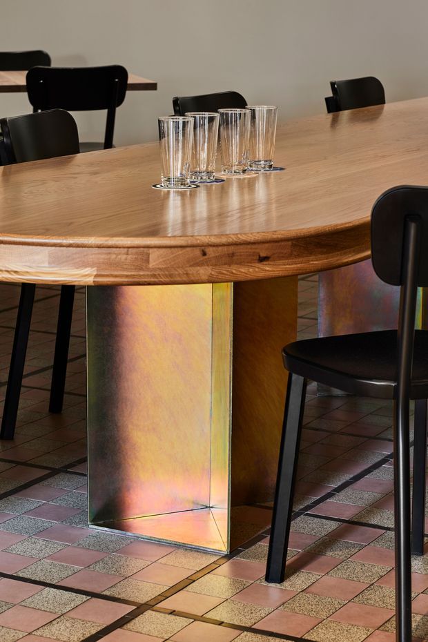 A close-up interior view of a dining area centred on a large round-edged timber dining table with an iridescent metal pedestal base. Black dining chairs surround the table, which holds a cluster of clear drinking glasses. The floor is finished in a geometric checkerboard pattern of muted pink, beige and dark tiles, creating a bold retro-inspired setting.