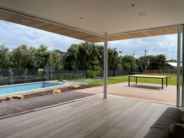 A contemporary covered outdoor entertaining area opening onto a timber deck, with a swimming pool, glass pool fencing, lawn, and a table-tennis table in the landscaped yard. The space has a broad flat ceiling with recessed downlights, timber-look soffit lining, slender support columns, and large open glazed sliding doors. A sofa is partially visible in the foreground.
