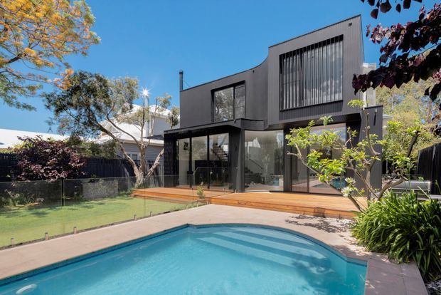 Exterior view of a contemporary two-storey dark-clad residence with large glazed openings, vertical window screening, and a timber deck overlooking a curved swimming pool. The landscaped suburban setting includes lawn, mature trees, boundary fencing, and bright clear daylight.