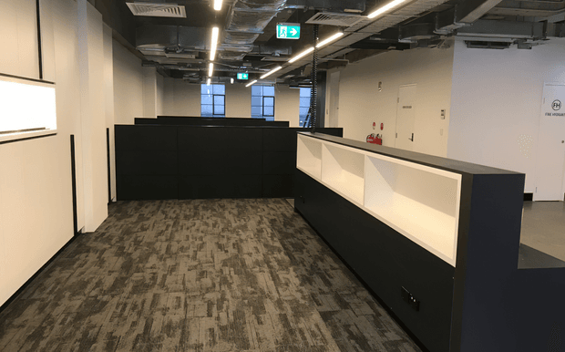 An empty contemporary commercial office interior featuring dark built-in partition walls and illuminated recessed display or reception-style panels. The space has patterned grey carpet flooring, white walls, exposed services and ductwork in the ceiling, linear strip lighting, emergency exit signage, and fire-safety equipment. The layout appears to be an open-plan office or circulation area with workstations or low partitions.