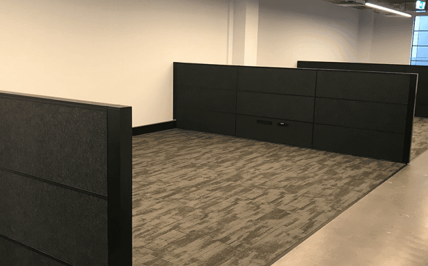 A modern open-plan commercial office interior with dark acoustic partition panels dividing carpeted work areas. The space has neutral painted walls, exposed linear ceiling lighting, and a polished concrete circulation path along the right side. No desks or loose furnishings are visible.