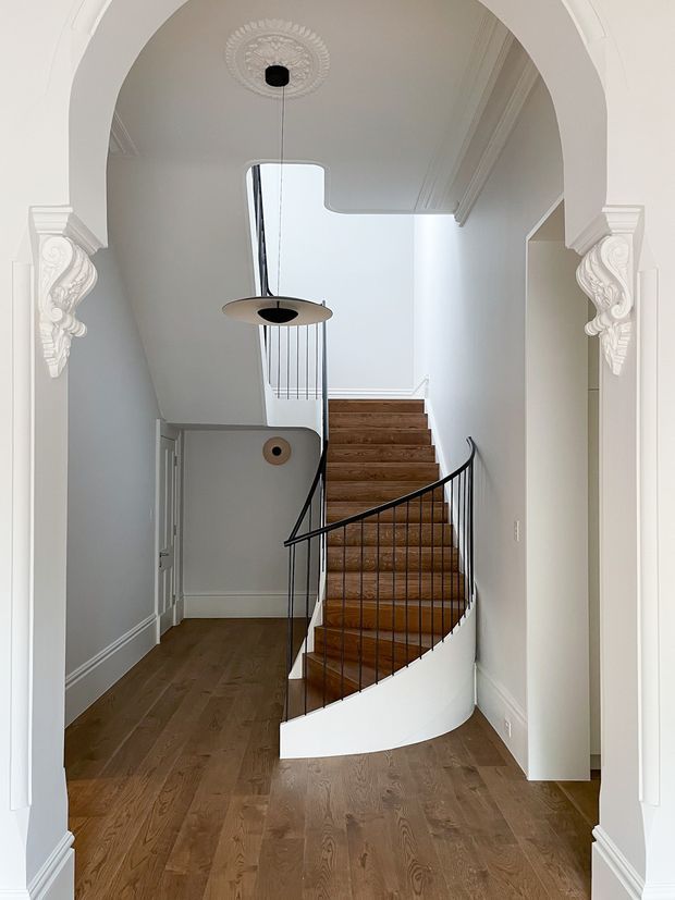 A bright, refined residential entry hall framed by a large white arched opening with ornate decorative plaster corbels and ceiling rosette. The space features warm timber flooring, a curved staircase with matching timber treads, white balustrade walls, and slim black metal railings. A sculptural pendant light hangs over the stairwell, while traditional wall mouldings and cornices contrast with the clean contemporary staircase detailing.
