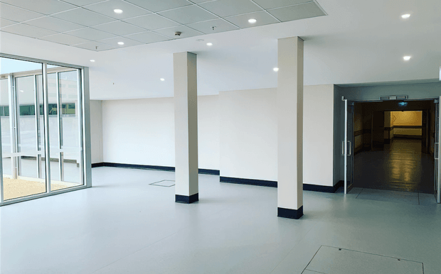 A bright, empty commercial interior or institutional lobby with a large open floor area, white walls, square structural columns, dark skirting, recessed ceiling lights, and a suspended grid ceiling. Full-height glazed doors and windows open to the left, while a dark-framed doorway leads to a dim corridor or service area on the right. The space has a clean, utilitarian fit-out.