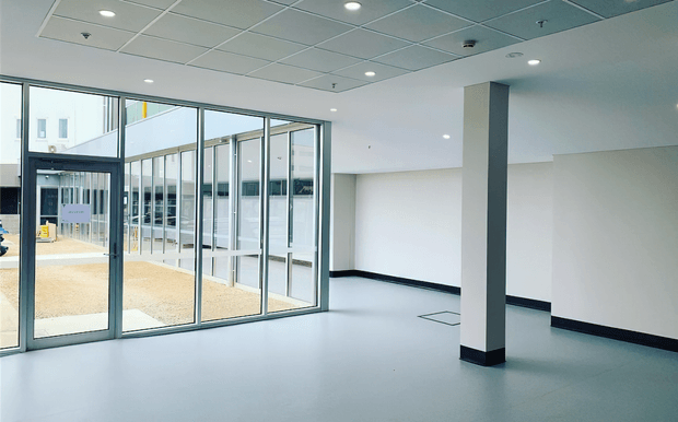 A bright, empty commercial interior or building entrance with a smooth light-grey floor, white walls, black skirting, exposed square column, suspended acoustic ceiling with recessed downlights, and extensive aluminium-framed floor-to-ceiling glazing and glazed entry doors. An exterior courtyard or covered walkway is visible beyond the glazing. The space is minimal and newly fitted out, with no furniture.