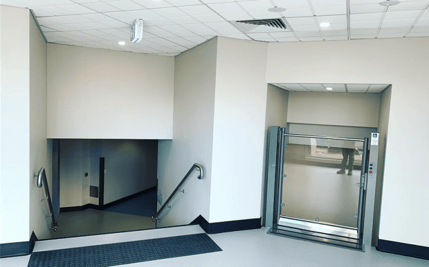 Bright, utilitarian interior entrance area with a descending stairwell on the left and a glass-fronted elevator on the right. The space has smooth off-white painted walls, dark trim and flooring at the stair landing, a suspended acoustic tile ceiling with recessed lighting and visible HVAC, stainless-steel handrails, and accessible lift access. The image appears to show a commercial or institutional building circulation zone.
