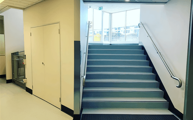 A utilitarian commercial or institutional interior showing a straight staircase leading up to a bright glazed landing. The stairs have dark blue-grey treads and risers with light edge strips, stainless-steel handrails on both sides, and pale walls with dark protective wall trim. A plain double door and service equipment are visible beside the stairwell. The space has a suspended acoustic ceiling and cool, functional finishes.
