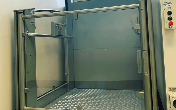 An enclosed commercial goods lift or service elevator with a diamond-plate metal floor, grey metal wall panels, stainless-steel framing, hinged access doors, and visible lift control panels. The utilitarian space is photographed from the doorway.