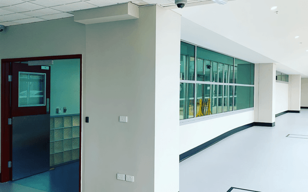 A clean institutional or commercial interior corridor with white walls, a pale smooth floor, dark skirting, a suspended grid ceiling, and large internal glazed windows looking into another workspace. A dark red-brown framed door with a small window and lower protective panel opens to a secondary room on the left. The space is bright, utilitarian, and minimally furnished.