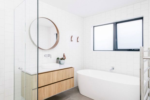 Bright contemporary bathroom with a freestanding white bathtub, frameless glass shower enclosure, floating timber vanity with integrated basin, round wall mirror, white tiled walls, dark grey floor tiles, black-framed window, and a heated towel rail. The space has a clean, minimal and neutral presentation.
