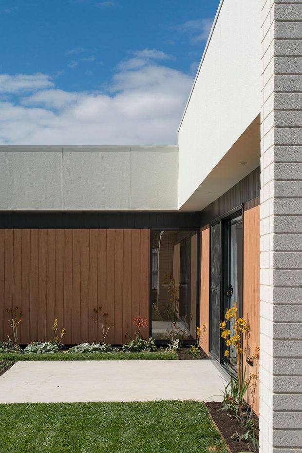 A modern residential courtyard viewed from outdoors, featuring crisp white rendered and light-grey brick walls, a projecting flat-roof form, dark-framed glazing, vertical timber wall cladding, and a simple concrete-paved path bordered by lawn and planted garden beds. The composition has clean geometric lines and a bright contemporary exterior aesthetic.