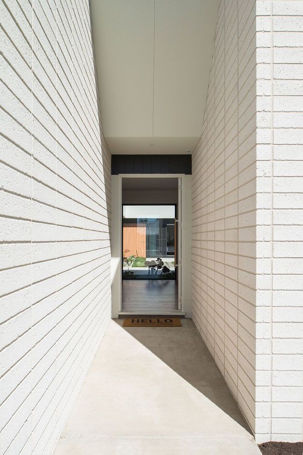 A narrow, covered entry passage flanked by white-painted brick walls, leading to a glazed front door. The recessed ceiling and concrete floor create a clean, minimalist approach, with a small doormat at the threshold. Through the glass, a bright interior and courtyard-like area are visible.