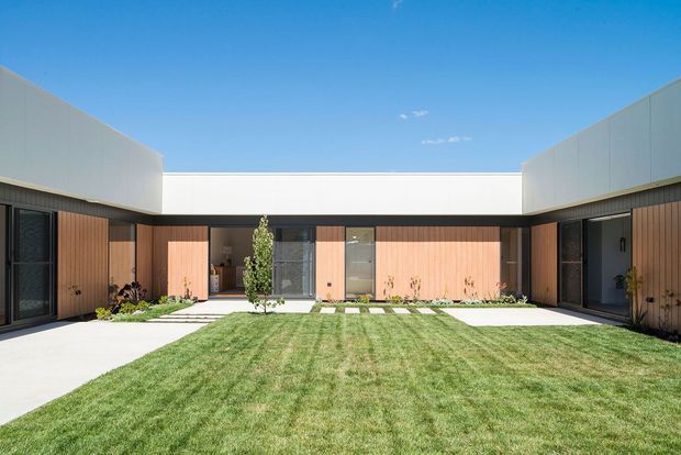 A low-slung contemporary courtyard house arranged in a U-shaped plan around a broad central lawn. The facade combines warm vertical timber cladding, dark-framed glazing and doors, crisp white upper volumes, and a dark horizontal roofline. A paved path crosses the landscaped courtyard, with small planted garden beds and a central young tree beneath a clear blue sky.