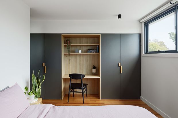 A compact, contemporary bedroom with a built-in dark charcoal storage wall and integrated timber study nook. The central open shelving and desk area is paired with a black chair, while a bed with pale pink bedding occupies the foreground. A large dark-framed awning window brings in natural light, and a small indoor plant adds greenery. The room has white walls, a simple white ceiling with a recessed black spotlight, and warm timber flooring.