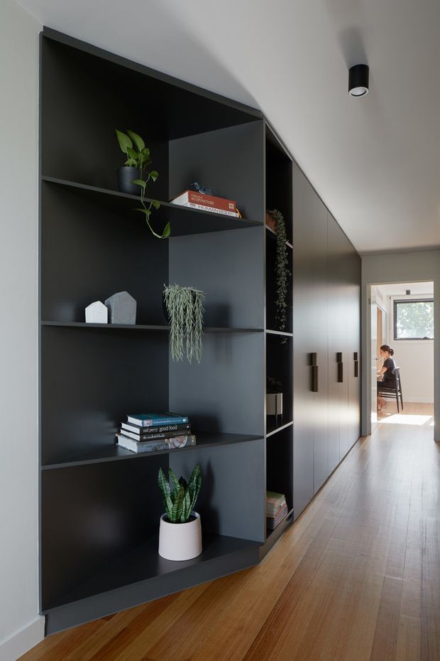 A contemporary hallway with a long, dark built-in shelving and cabinetry wall. Open niches display potted plants, books, and small decorative objects, while closed cupboards continue toward a bright room at the end of the corridor. The space has warm timber flooring, pale walls, minimalist black cylindrical track lighting, and clean-lined joinery.