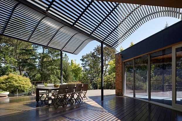 A spacious outdoor timber deck with a dining table and chairs beside a glazed brick-and-dark-clad building. A dramatic curved slatted pergola creates strong striped shadows across the deck, while surrounding mature greenery is visible beyond the railing.