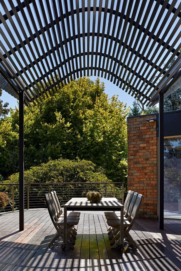 A sunlit outdoor dining deck beneath a dramatic curved pergola made of closely spaced dark timber slats. A rectangular dining table with folding chairs sits on timber decking, with a slim metal cable balustrade and dense garden greenery beyond. A red-brown brick wall and dark-framed glazing define the side of the adjoining building. Strong striped shadows create a graphic, contemporary architectural setting.