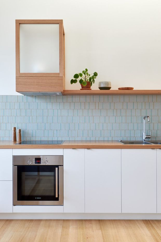 A bright, compact contemporary kitchen with white flat-panel cabinetry, a timber benchtop and matching timber floor, pale blue vertically arranged tiled splashback, stainless-steel oven and sink, and a timber-framed wall cabinet with open display shelf. Minimal ceramic vessels and a small plant sit on the shelf.