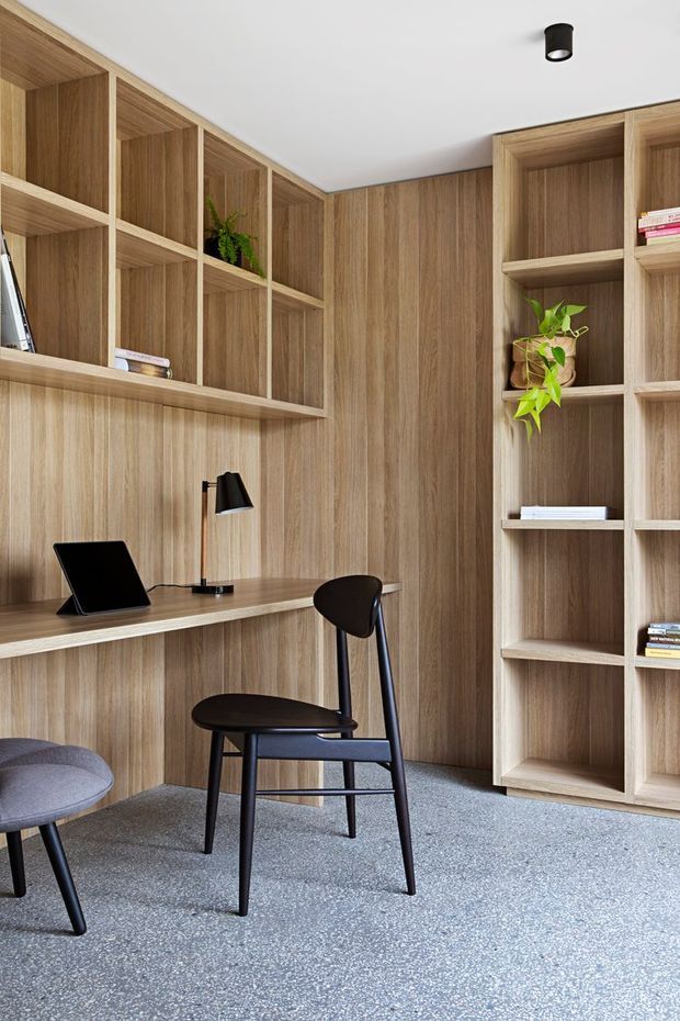A compact, built-in home office with extensive light timber wall panelling, integrated open shelving, and a floating desk. A black laptop and desk lamp sit on the workspace, with a dark timber-and-black chair and a grey upholstered stool. The room has a white ceiling with a small black cylindrical spotlight and a grey speckled floor covering. Minimal, warm and highly integrated joinery dominates the composition.