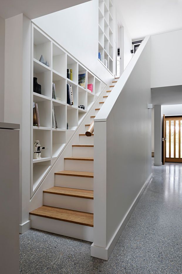 Bright modern interior hallway centred on a staircase with white painted walls and integrated open shelving along the stair wall. The stair treads are warm timber, while the surrounding floor is a polished grey terrazzo-like finish. A white balustrade and half-height partition frame the stairs, with a timber-framed glazed entry door visible in the background.