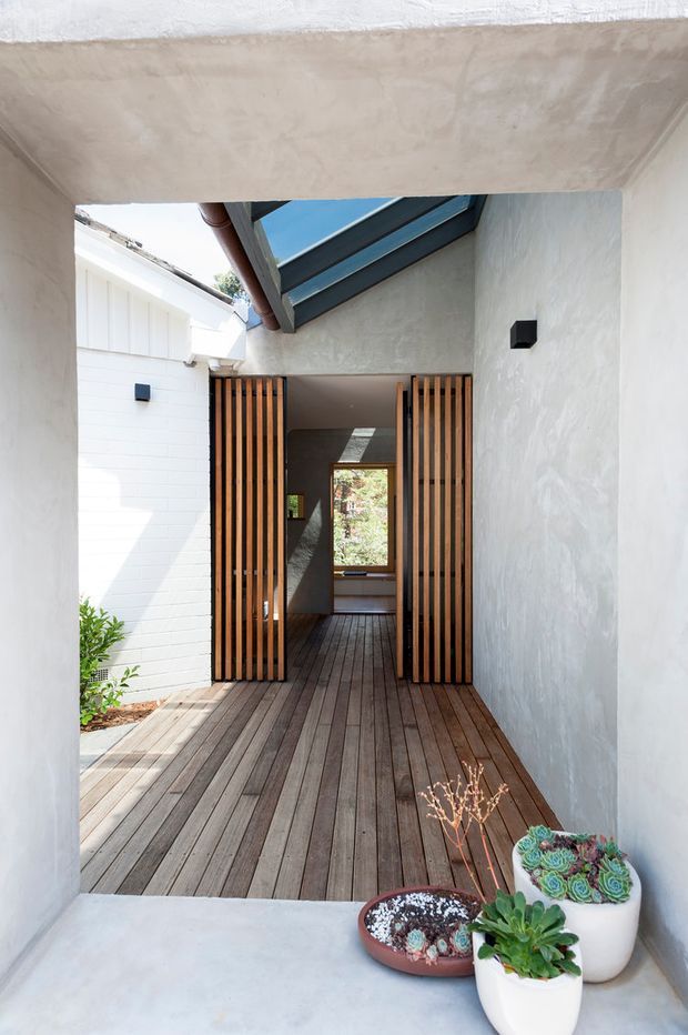 A narrow, sheltered residential entry courtyard viewed from beneath a concrete canopy. The space features white textured masonry and rendered walls, a dark timber deck, paired vertical timber slat gates, a glazed rooflight/skylight, black wall lights, and potted succulents. The deck leads through the timber gates toward a bright interior or rear courtyard, creating a calm, modern indoor-outdoor transition.