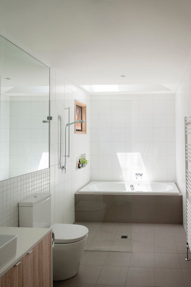Bright contemporary bathroom with a full-width tiled shower area, freestanding toilet, vanity, wall-mounted towel warmer, and built-in bathtub beneath a large skylight. White tiled walls and pale floor tiles create a clean, airy interior, with timber-framed window detailing and dark bathtub cabinetry providing contrast.