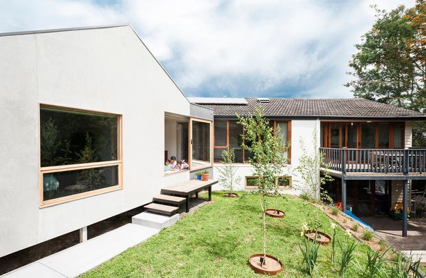A contemporary single-storey residential extension arranged around a small landscaped courtyard. The pale rendered addition has large timber-framed windows and a simple sloping roofline, connected to an older-looking tiled-roof house with a raised timber deck and dark balustrade. Concrete steps and paths lead through the grass courtyard, with established trees and garden planting visible around the building.