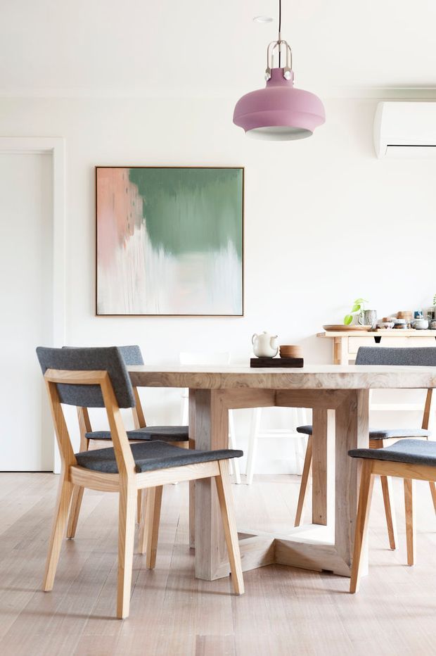 Bright, minimalist dining area with a solid light timber dining table and matching timber-framed chairs upholstered in dark grey fabric. White walls and pale timber flooring create an airy backdrop, complemented by a large abstract pastel painting, a mauve pendant light, and a compact kitchen or servery visible behind the table.