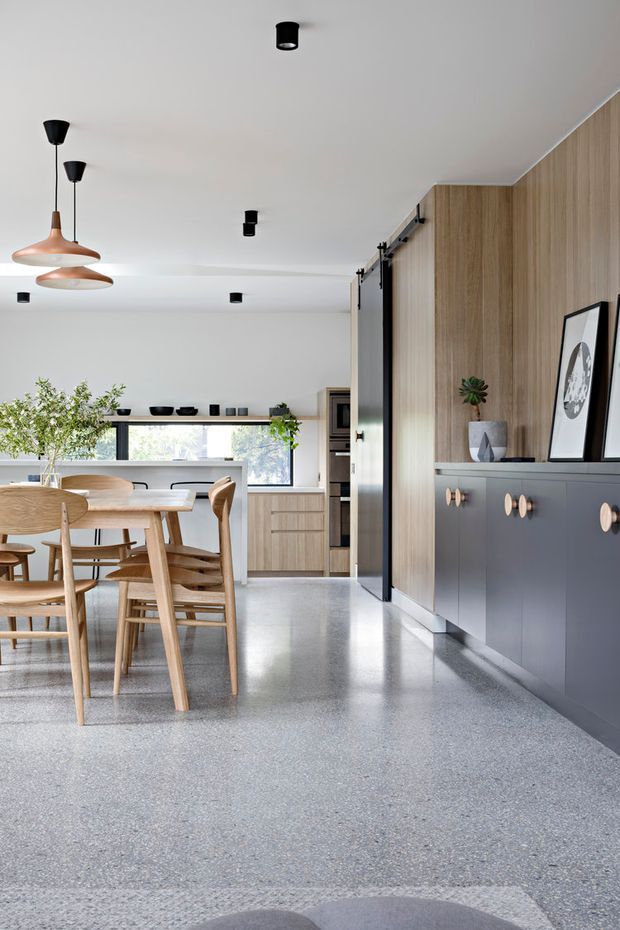 Open-plan residential dining and kitchen area with a warm, contemporary fit-out. A light timber dining table and chairs sit beside a compact kitchen, while the right wall features full-height vertical timber panelling above dark grey built-in cabinetry with round timber knobs. The space has a smooth pale grey polished floor, white ceiling and walls, black track and pendant lighting, large windows, and sliding dark-framed doors.