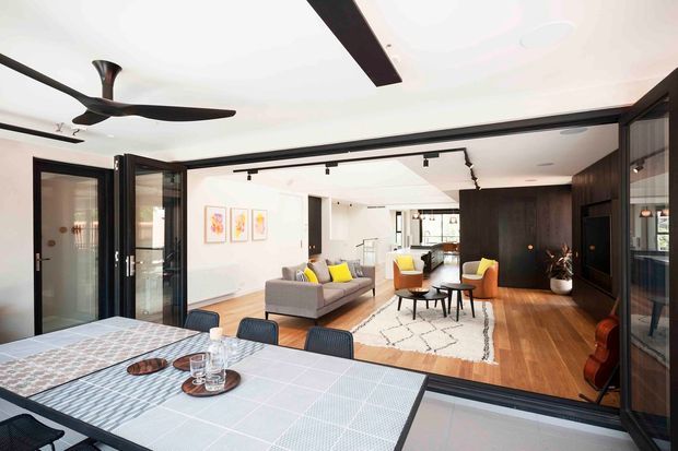 Open-plan contemporary interior viewed from a dining area through fully opened black-framed glazed doors. The foreground contains a dining table, while the adjoining living room features a grey sofa, yellow accent cushions, lounge chairs, coffee tables, a patterned rug, timber flooring, dark built-in cabinetry, track lighting, and a ceiling fan. The space is bright and connected through wide indoor-outdoor-style openings.